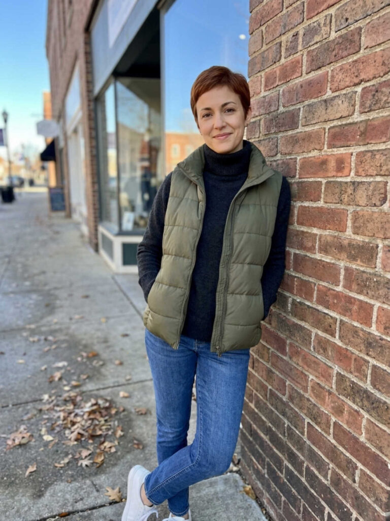 Woman in a turtleneck and puffer vest leaning against a brick wall in winter.