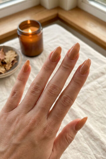 A hand with warm caramel nude almond nails resting on a linen tablecloth.