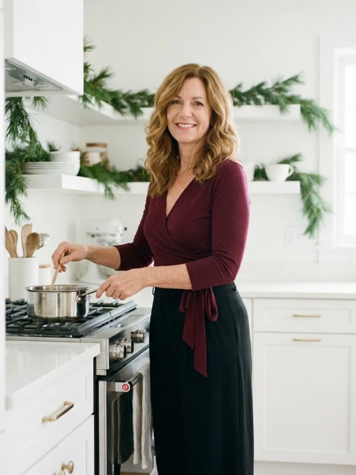 Woman in a burgundy wrap top and black wide-leg pants stirring a pot in a bright kitchen decorated with a pine garland.