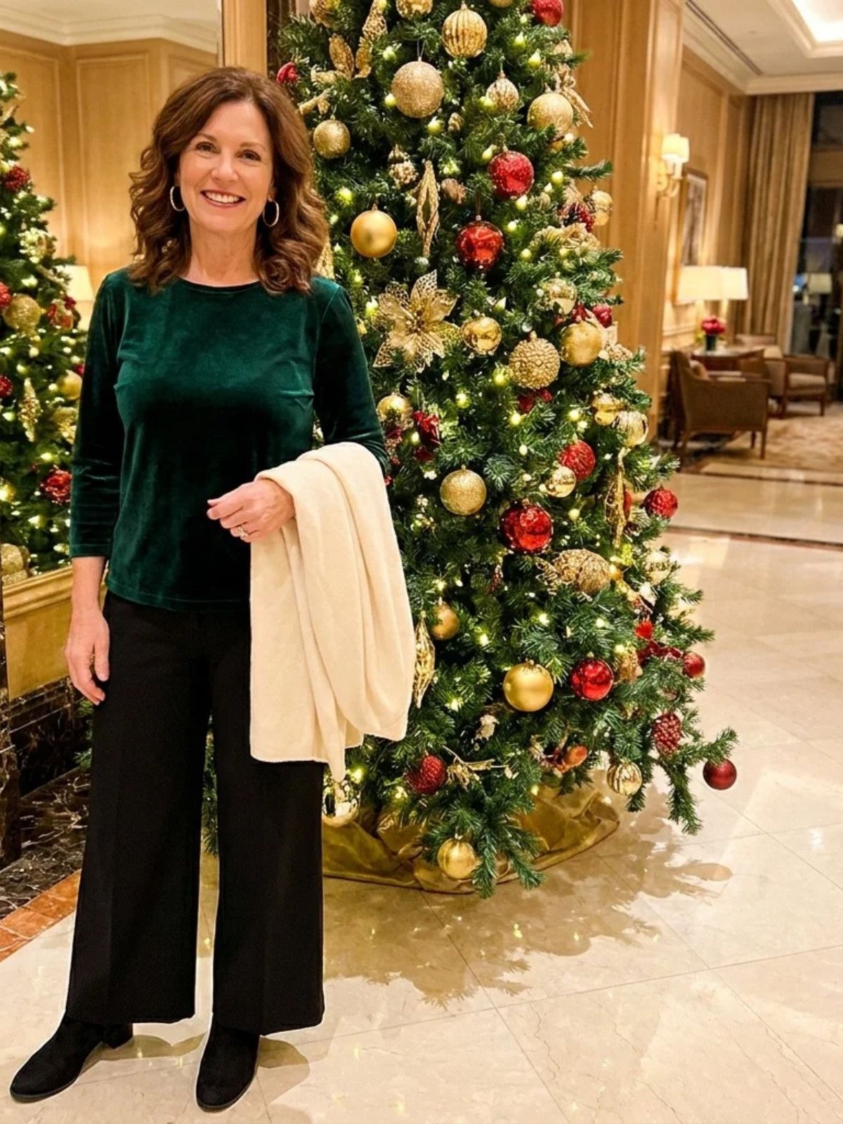 A woman in an emerald velvet top and black wide-leg pants stands in a resort lobby beside a large decorated Christmas tree.