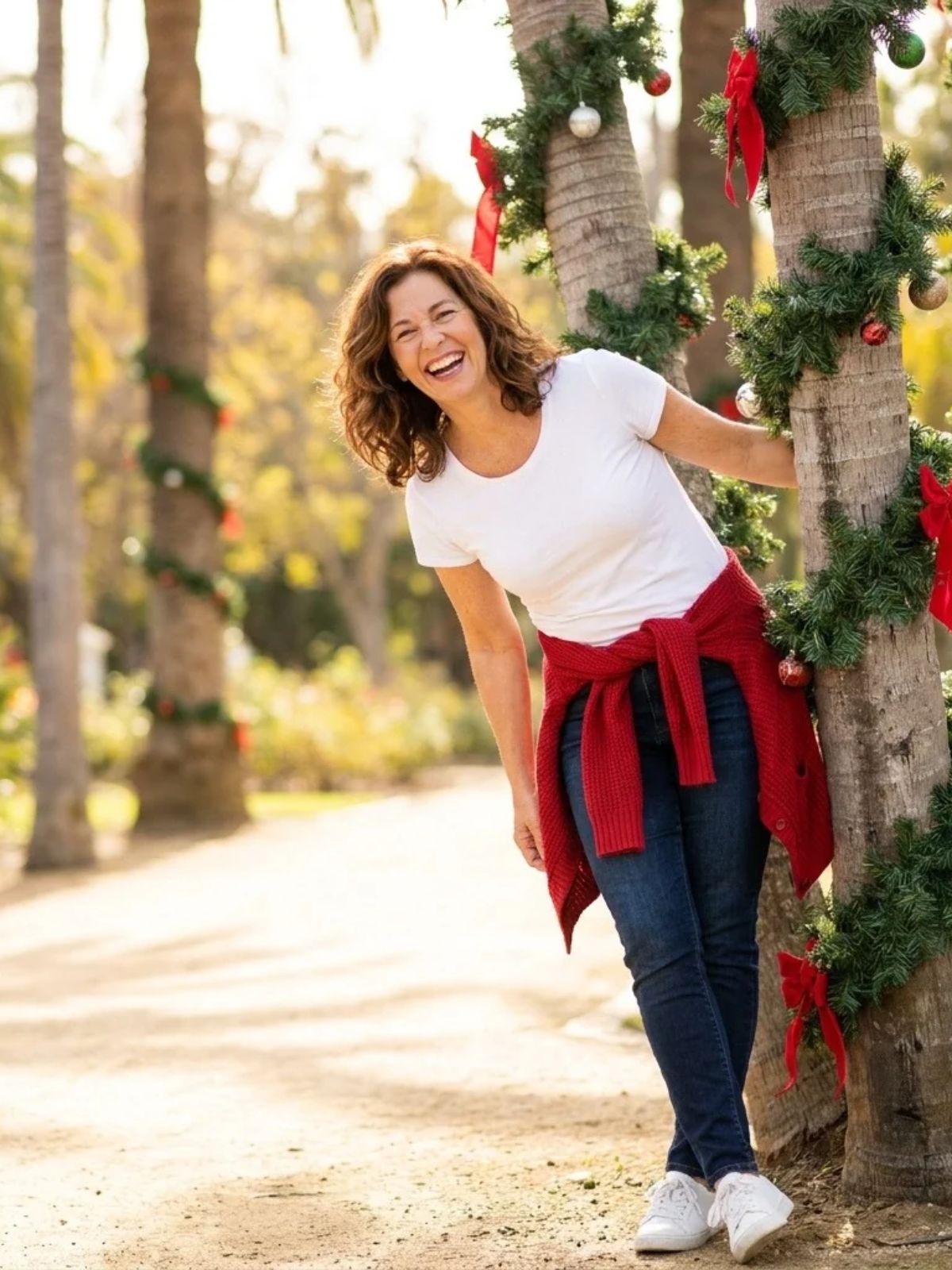  A woman in a white tee ties a red cardigan around her waist on a sunny afternoon beside palm trees wrapped in holiday garlands.