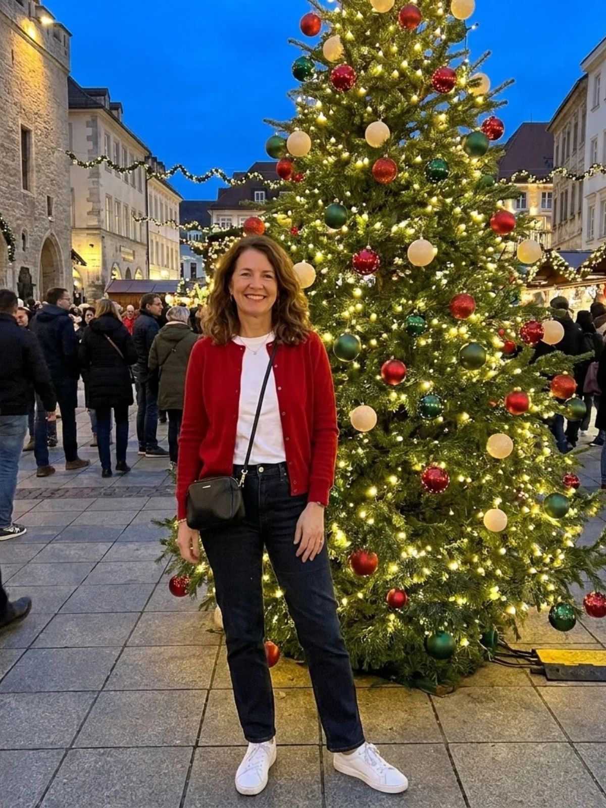 A woman in a red cardigan over a white tee and dark straight-leg jeans smiles beside a tall lit Christmas tree.