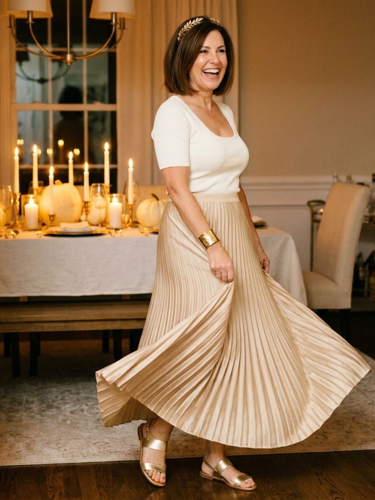Woman in a gold pleated maxi skirt, ivory top, and gold leaf headband turning in a candlelit dining room.