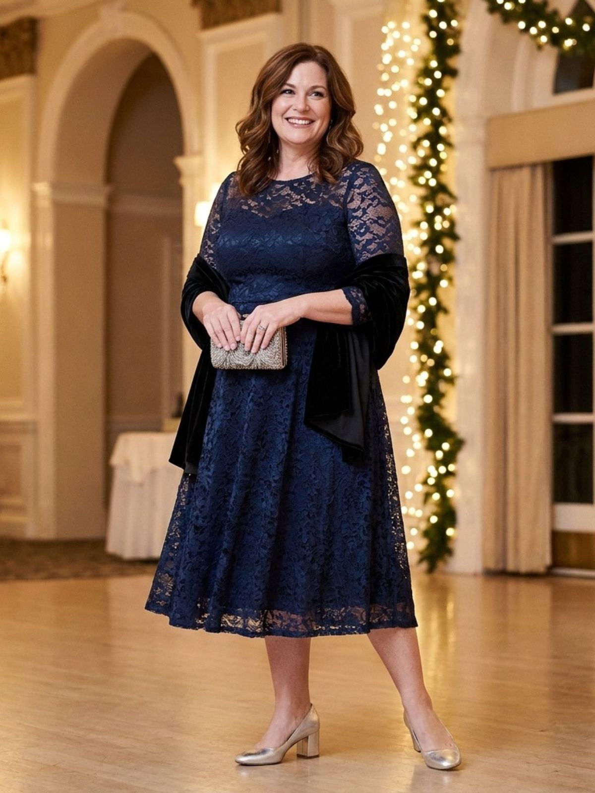  A woman in a midnight blue lace fit-and-flare dress with sheer three-quarter sleeves, silver block heels and a beaded clutch in a ballroom.