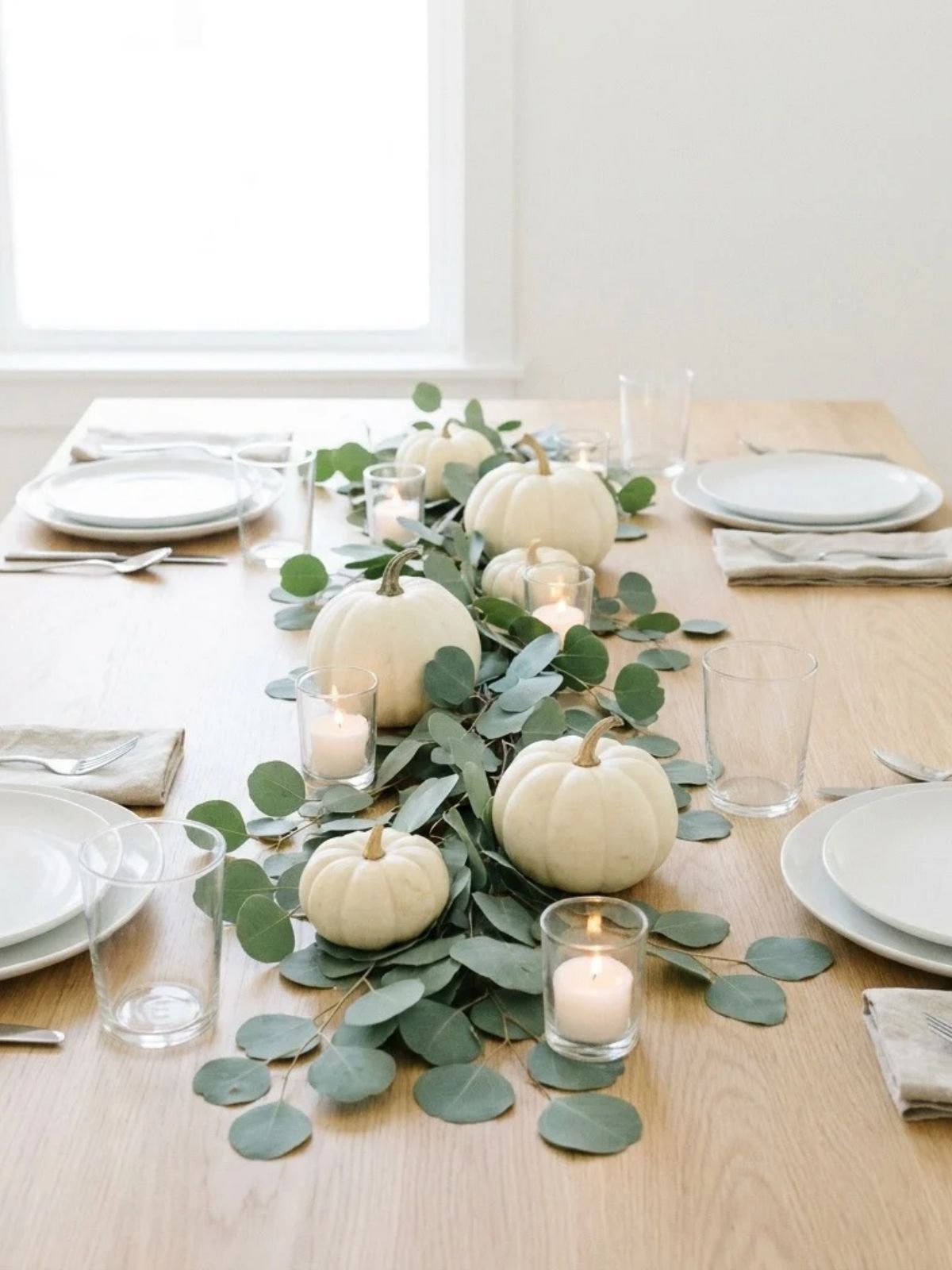 modern Thanksgiving table with white pumpkins, silver dollar eucalyptus, white dishes, and glass votives on a light oak table.