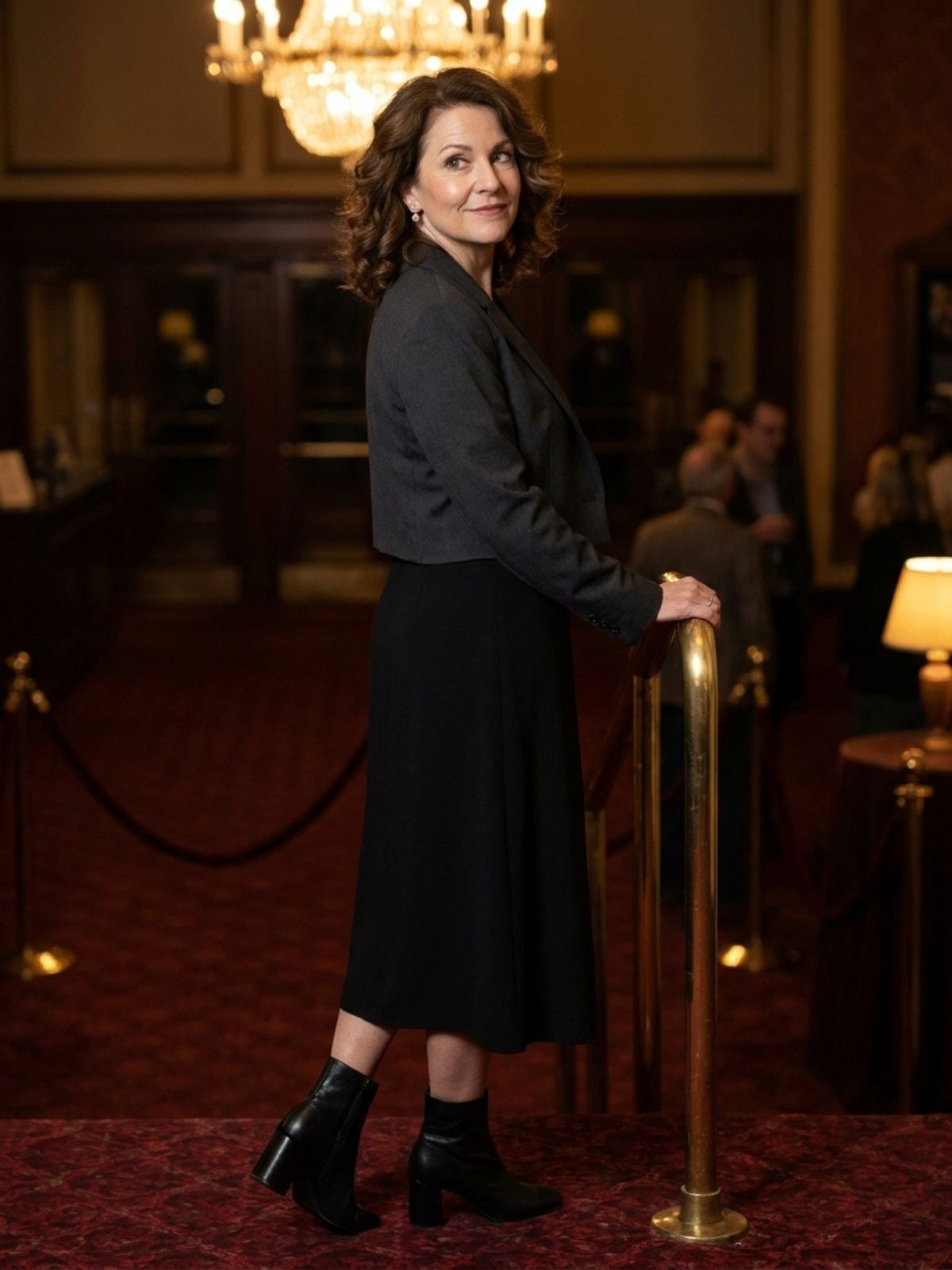 Woman in a black wrap dress, cropped charcoal blazer, and ankle boots on a theater lobby staircase.